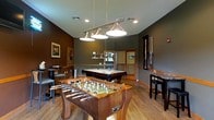 This single-floor recreational space is thoughtfully designed as a dedicated game room, offering an inviting atmosphere for entertainment and socializing. The room is anchored by a classic pool table positioned at the center, illuminated by a set of stylish pendant lights with metallic shades that hang overhead, casting focused light across the playing surface. A solid wood foosball table takes prominence in the foreground, adding a dynamic focal point to the room's layout. Bar-height tables and stools are arranged along the walls, providing comfortable seating for spectators and guests. The walls are finished in deep, warm tones of gray and olive, contributing to the room's relaxed yet polished ambiance. Light hardwood flooring runs throughout, adding warmth and contrast to the darker wall palette. A cue rack mounted on the wall keeps equipment neatly organized, while framed artwork adds a decorative touch. Recessed lighting complements the pendant fixtures, ensuring the space is well-lit throughout. A doorway visible in the background suggests connectivity to adjacent areas, and a window allows for a degree of natural light to filter in, completing this well-balanced and functional recreational environment.