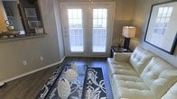 This single-story space showcases a well-appointed living area that combines modern design with a warm, inviting ambiance. The room is anchored by rich, dark hardwood flooring that runs throughout, providing a sleek and polished foundation for the space. A standout feature is the set of white French doors, which serve as the focal point of the room, fitted with horizontal blinds that allow for controlled natural light. The doors invite an abundance of daylight into the space, enhancing its open and airy feel. The walls are finished in a contemporary warm gray tone, complementing the dark flooring and contributing to a cohesive, neutral color palette. A tufted white leather sofa adds a touch of elegance, paired with a glass-top coffee table adorned with decorative sculptural pieces. A bold black-and-white patterned area rug grounds the seating arrangement and adds a graphic design element to the room. A dark side table with a classic lamp provides both functional lighting and a stylistic accent. The overall aesthetic of the space is modern and refined, with thoughtful attention to contrast and detail throughout.