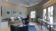 This single-floor space is thoughtfully designed to serve as a functional and welcoming business center or leasing office. The interior is finished with neutral warm-toned walls, crown molding, and light-colored flooring, creating a polished and professional ambiance throughout.

A dedicated computer workstation area lines one wall, equipped with multiple desktop computers and ergonomic chairs, making it well-suited for productivity and research. A ceiling fan with a light fixture anchors the center of the room, providing both air circulation and supplemental lighting alongside recessed ceiling lights that illuminate the space evenly.

Natural light filters through a large window dressed with elegant floor-length drapes, offering a view of lush greenery outside. Beneath the window, a compact white storage unit adds practical organization to the space. A comfortable black leather accent chair and a navy area rug define a casual seating zone, ideal for informal meetings or relaxed conversation.

On the opposite side, a high bar-style table paired with sleek modern chairs provides additional workspace or a spot for collaborative discussions. Contemporary abstract artwork adorns the walls, adding a touch of color and personality to the otherwise understated décor. Overall, the space balances functionality with comfort in a clean, modern aesthetic.