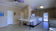 This single-floor communal laundry facility is designed with both functionality and comfort in mind. The space features vaulted ceilings that lend an open, airy feel, complemented by multiple ceiling fans that ensure consistent air circulation throughout. The walls are finished in a warm neutral tone, trimmed with clean white crown molding and chair rail accents that add a touch of polish to the utilitarian space.

A row of top-load washing machines and dryers lines the far wall, positioned beneath a window that allows natural light to filter in, brightening the workspace. A sleek white folding table with distinctive hairpin legs sits centrally in the room, providing ample space for sorting and folding laundry. The flooring is a smooth, easy-to-maintain surface suited for high-traffic use.

Safety is addressed with a mounted fire extinguisher on the wall, and multiple ceiling-mounted light fixtures ensure the room is well-lit at all times. A private door leads to an adjacent room or utility area, adding to the overall convenience of the space. The layout is thoughtfully organized to allow multiple users to move freely and comfortably, making this laundry facility a practical and welcoming amenity.
