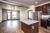 This contemporary single-floor unit combines modern industrial design with practical living. The open-concept layout connects the kitchen and living area seamlessly, anchored by rich hardwood-style flooring throughout. A defining architectural feature is the exposed silver ductwork running overhead, adding an industrial loft character to the space.

The kitchen is well-appointed with dark espresso cabinetry, light quartz-style countertops, a full suite of stainless steel appliances including a refrigerator, range, microwave, and dishwasher, as well as a central island for additional prep space and storage. Open shelving on the wall provides extra display and storage options. A sliding barn door adds a stylish, space-saving touch near the kitchen entry.

The living area is bright and airy, with large windows and a glass-panel entry door drawing in generous natural light and offering outdoor views. Ceiling fans are present throughout, ensuring comfortable air circulation. The bedroom is spacious, featuring its own ceiling fan and large window, and connects directly to a bathroom and a dedicated walk-in closet with built-in shelving. A second bathroom with a vanity and mirror is also present. A utility area houses the water heater and electrical panel, keeping essential systems conveniently accessible.