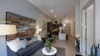 This thoughtfully designed single-floor residence showcases a seamless open-concept layout that effortlessly connects the living, dining, and kitchen areas into one cohesive and inviting space. The living area is anchored by a plush charcoal sofa, complemented by tasteful décor including a large statement artwork in warm earth tones and an assortment of decorative wall accents. Recessed lighting and soaring vaulted ceilings enhance the sense of openness and volume throughout.

The kitchen is well-appointed with dark cabinetry, stainless steel appliances, and a bright white breakfast bar that offers additional seating and serves as a natural dividing element between the cooking and living spaces. A pendant light fixture overhead adds a refined touch to this functional area. Adjacent to the kitchen, the dining space accommodates a full table and chairs, making it ideal for everyday meals or entertaining.

Warm-toned luxury vinyl plank flooring runs throughout the main living areas, providing both durability and a contemporary aesthetic. Neutral gray walls create a versatile backdrop that complements the modern furnishings. Natural light filters in, contributing to the bright and welcoming ambiance. A private entry door at the rear of the hallway completes the layout, offering convenience and a sense of separation from shared spaces.