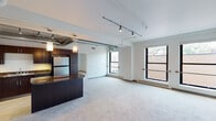 This single-floor loft-style unit blends industrial and contemporary design elements throughout. The open-plan living and dining area is defined by soaring ceilings with exposed spiral ductwork and sleek track lighting, lending the space a distinctly urban character. Large, black-framed windows flood the main living area with abundant natural light while offering views of the surrounding cityscape. Plush wall-to-wall carpeting adds warmth and comfort underfoot across the expansive living space.

The kitchen is seamlessly integrated into the open layout via a dark-toned breakfast bar with rich granite countertops. Cabinetry in a deep espresso finish pairs with stainless steel appliances — including a refrigerator, built-in microwave, and dishwasher — to create a functional and stylish cooking space.

The bedroom is a well-proportioned, carpeted retreat featuring a large window with a baseboard heating unit below and a frosted glass door that adds a modern touch while allowing light to pass through. The full bathroom is finished with a dark wood vanity, crisp white countertop, hardwood-style flooring, and a full tub-shower combination, offering a clean and practical design.