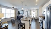This single-floor home showcases an open-concept layout that seamlessly connects the kitchen, dining, and living areas into one cohesive, flowing space. The kitchen serves as the central hub, anchored by a granite-topped island with an undermount sink, pendant lighting, and a built-in dishwasher, complemented by a bar stool seating for casual dining. White cabinetry adds a clean, bright contrast to the rich, dark hardwood flooring that extends throughout the entire space.

The dining area sits adjacent to the kitchen, bathed in natural light streaming through large windows dressed with elegant floral-patterned curtains. A round dining table with upholstered chairs creates an inviting setting for meals.

The living area is comfortably appointed with a wall-mounted flat-screen television, a dark media console, and a ceiling fan that enhances airflow and comfort. Multiple windows fitted with blinds and layered curtains allow for flexible light control while maintaining the space's bright, airy ambiance. The cohesive neutral color palette and consistent flooring unify each zone, creating a welcoming and functional single-level home.