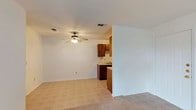 This single-story space offers a practical, open-concept layout that visually connects the living area to the kitchen. The living area is finished with neutral-toned carpet and painted in crisp white, creating a clean and versatile backdrop. A ceiling fan with an integrated light fixture is centrally positioned, providing both air circulation and ambient lighting.

A partial wall divides the living space from the kitchen, maintaining an open feel while subtly defining each area. The kitchen features medium-toned wood cabinetry along the upper and lower sections, offering ample storage. Light-colored countertops provide workspace, and the flooring transitions to tile, a practical choice for the kitchen zone. A sink is visible along the counter, and the cabinetry layout suggests efficient use of the available space.

HVAC vents are visible in the ceiling throughout, indicating a central air system. The neutral color palette and straightforward design make the space highly adaptable. Overall, the layout prioritizes functionality and ease of movement between the living and kitchen areas.