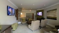 This thoughtfully designed lobby lounge offers a refined and comfortable environment that makes an immediate impression upon entry. The space features a neutral color palette accented by rich olive green walls and a textured wood-panel feature wall, creating a warm and contemporary aesthetic. Recessed ceiling lighting, complemented by a soft pendant lamp, provides layered illumination throughout the room. Seating is arranged in a relaxed, conversational layout with plush upholstered armchairs in muted tones and coordinating green accent cushions, offering a comfortable place to sit and unwind. Square ottomans in a sage green finish provide additional flexible seating options. A wall-mounted flat-screen television is positioned on the olive-toned accent wall, adding a modern entertainment element to the lounge. Decorative touches, including sculptural branch arrangements and wall art, contribute to the space's polished character. Elevator access is visible from the lounge, integrating practical functionality seamlessly into the design. The carpeted flooring and crown molding details further enhance the overall sense of quality and attention to detail, making this lobby a welcoming gateway to the building.