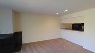 This single-floor space features an open and straightforward layout designed for functional living. The main living area is finished with wall-to-wall carpeting in a neutral tan tone, contributing to a warm and understated ambiance throughout. The walls are painted in a soft cream color, creating a clean and bright backdrop that complements the overall simplicity of the space.

Recessed lighting is installed in the ceiling, providing even and adequate illumination across the room. The ceiling itself is smooth and finished in white, maintaining a cohesive and tidy appearance.

Toward the back of the space, a partial wall separates the living area from a kitchen nook. The kitchen features a countertop with a granite-style surface, and upper cabinetry finished in white is visible above. A refrigerator can be seen tucked into the kitchen area, indicating a functional setup despite the compact footprint.

Electrical outlets are positioned along the walls at standard intervals, and the overall condition of the space appears well-maintained. The open feel of the main room offers flexibility for various furniture arrangements, making it a versatile and practical space.