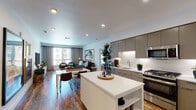 This single-floor residence is thoughtfully designed with a contemporary aesthetic and functional open-concept layout. The kitchen is a standout feature, equipped with flat-front taupe cabinetry extending to the ceiling, white quartz countertops, stainless steel appliances including a gas range, built-in microwave, and French door refrigerator. A freestanding white island provides additional prep and storage space, while hardwood flooring and under-cabinet lighting enhance the modern feel. The kitchen flows seamlessly into the dining and living areas, where large windows invite natural light and offer views of the surrounding neighborhood.

The living room is a comfortable, well-proportioned space anchored by a generous sofa seating arrangement and large-format wall art. The two bedrooms each feature plush carpeting or hardwood flooring, windows with urban views, and ample room for full bedroom furniture sets. One bedroom showcases a textured accent wall, adding warmth and depth.

The bathrooms offer distinct designs: one features a dual-sink vanity with wood cabinetry and bold deep-blue walls, while the second includes a soaking tub with a curved shower rod and neutral tile surround, providing a clean, spa-inspired atmosphere throughout.