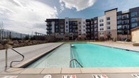 This outdoor amenity space in Sandy, Utah, is designed to offer a comprehensive recreational experience within a modern residential community. The centerpiece is a generously sized swimming pool with clear, blue-tinted water, graded from a shallow entry point to a deeper end, surrounded by a wide, clean concrete pool deck ideal for lounging. The pool area is framed by natural stone landscaping and black metal fencing, which contributes to both the aesthetic and safety of the space.

Adjacent to the pool, a covered patio area features large-format concrete pavers, sturdy dark wood posts, and an outdoor grilling station, making it well-suited for outdoor gatherings. The patio offers open sightlines toward the surrounding mountains, providing a scenic backdrop that enhances the outdoor experience.

The fitness center is accessible directly from the pool deck through wide glass sliding doors, offering transparency and visual connectivity between the indoor and outdoor spaces. The gym interior features carpet flooring, bold graphic signage, and visible exercise equipment. The building exterior incorporates natural stone cladding and large windows, lending a contemporary yet grounded aesthetic that complements the Utah landscape.