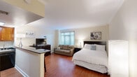 This single-floor unit in New Orleans, Louisiana, offers a well-organized layout that maximizes its compact footprint. The main living and sleeping area is an open, versatile space finished with warm hardwood flooring and crown molding throughout, lending a polished, classic character to the interior. A large window allows natural light to brighten the room, which comfortably accommodates a queen bed, sofa, and entertainment setup, illustrating the efficient use of the available space.

The kitchen is separated from the main area by a peninsula breakfast bar topped with dark granite countertops, providing both a visual boundary and casual dining seating with bar stools. Rich wood cabinetry extends to the ceiling, offering generous storage, while black appliances including a full-size refrigerator, gas range, and microwave complete the functional cooking space.

A hallway connects the main area to additional sleeping quarters, where hardwood floors continue and the clean, neutral palette is maintained. Crown molding and white baseboards are consistent throughout, reinforcing a cohesive, tidy aesthetic. The overall ambiance is understated and practical, suited to those seeking a comfortable, well-appointed space in the heart of New Orleans.