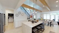 This three-storey home showcases a striking open-concept main level anchored by a spacious kitchen with a large waterfall-edge marble island topped in rich white-and-grey veining. The island comfortably accommodates bar seating and serves as the visual centerpiece of the space. Overhead, a cluster of gold-toned pendant lights adds warmth and a refined touch to the contemporary aesthetic.

Exposed light wood beams span the soaring vaulted ceiling, creating a sense of grandeur while lending a natural, organic contrast to the crisp white walls and light hardwood flooring found throughout. A modern staircase with sleek black metal balusters and a wood handrail connects the floors, rising dramatically through the open volume of the main living area.

The kitchen flows seamlessly into the living area, which is bathed in natural light from generous windows. White cabinetry with ample drawer storage lines the kitchen perimeter, keeping the space clean and functional. Geometric wall décor and a colorful abstract painting introduce personality without disrupting the space's modern sensibility. The overall ambiance is bright, airy, and thoughtfully designed.