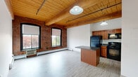 Located at 45 Willow St in Springfield, MA, this one-bedroom, one-bathroom unit blends historic industrial character with modern convenience. The open-concept living and kitchen area is the centerpiece of the space, featuring striking exposed brick walls, warm wood-planked ceilings supported by substantial timber beams, and light wood-style flooring. Large arched and rectangular black-framed windows line the brick walls, flooding the space with natural light and offering views of the surrounding neighborhood. The kitchen is well-appointed with rich wood cabinetry, dark granite-style countertops, black appliances, and a freestanding island that provides additional prep and dining space. Pendant lights hang from the beamed ceiling, adding warmth and a loft-inspired aesthetic. The bedroom continues the industrial character with the same exposed wood ceiling and beams, while plush carpeting adds comfort underfoot. Multiple closets throughout the unit offer generous storage. The full bathroom features a tub-shower combination, a wood-vanity with an integrated sink, and clean, bright finishes. With three closets spread throughout, storage is a practical highlight of this thoughtfully designed space.