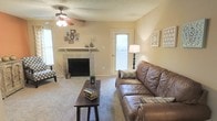 The living room of this single-story space is designed with comfort and functionality in mind. The room centers around a tile-surround fireplace with a clean white mantel, serving as a natural focal point and adding warmth to the overall atmosphere. Soft, neutral-toned carpet covers the floor, contributing to a relaxed and inviting ambiance. The ceiling features a textured finish and is equipped with a ceiling fan fitted with a light kit, providing both airflow and ambient lighting. Natural light enters the room through multiple windows, including a side-panel door window, brightening the warm beige walls. The layout accommodates generous seating, as evidenced by the placement of a leather sofa and accent chair. A rectangular wooden coffee table anchors the seating arrangement, offering a practical surface in the center of the room. Wall décor, including framed photos and decorative panels, adds a personalized character to the space. The overall design reflects a straightforward, livable layout well-suited for everyday use.