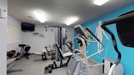 This fitness center is a thoughtfully designed, single-floor space dedicated to health and wellness. The room is finished with neutral gray walls, warm wood baseboards and door trim, and durable carpet flooring, creating a practical yet inviting environment for exercise.

The space is divided into distinct functional zones. A cardio area houses multiple treadmills, elliptical trainers, and a stationary spin bike, all arranged to allow comfortable use. A wall-mounted flat-screen television and a wall clock are positioned for easy viewing during workouts, while a window with blinds provides a connection to natural light. Storage cabinetry is built into one wall, offering convenient space for supplies.

The strength training zone is anchored by a Precor multi-station cable machine and a weight bench, providing a full range of resistance exercise options. Dumbbells are also available for free-weight training. A vibrant teal accent wall featuring bold silhouettes of athletes in motion adds energy and visual interest to this area, complemented by motivational text.

Overhead, flush-mounted panel lighting ensures the entire space is bright and well-illuminated. A glass-paneled door with a push bar leads to an adjacent area, adding to the accessibility of the facility. The overall ambiance is clean, modern, and purposefully organized to support a complete fitness routine.