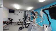 This fitness center is a thoughtfully designed, single-floor space dedicated to health and wellness. The room is finished with neutral gray walls, warm wood baseboards and door trim, and durable carpet flooring, creating a practical yet inviting environment for exercise.

The space is divided into distinct functional zones. A cardio area houses multiple treadmills, elliptical trainers, and a stationary spin bike, all arranged to allow comfortable use. A wall-mounted flat-screen television and a wall clock are positioned for easy viewing during workouts, while a window with blinds provides a connection to natural light. Storage cabinetry is built into one wall, offering convenient space for supplies.

The strength training zone is anchored by a Precor multi-station cable machine and a weight bench, providing a full range of resistance exercise options. Dumbbells are also available for free-weight training. A vibrant teal accent wall featuring bold silhouettes of athletes in motion adds energy and visual interest to this area, complemented by motivational text.

Overhead, flush-mounted panel lighting ensures the entire space is bright and well-illuminated. A glass-paneled door with a push bar leads to an adjacent area, adding to the accessibility of the facility. The overall ambiance is clean, modern, and purposefully organized to support a complete fitness routine.