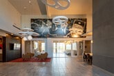 The Kennedy's leasing lobby is a visually dynamic, single-floor space that makes an immediate impression through its soaring ceilings and expansive open floor plan. Large-format tile flooring extends throughout the main area, lending a clean and polished aesthetic. Dramatic statement lighting—including sculptural artichoke pendants and interlocking ring chandeliers—hangs from the elevated ceiling, creating striking focal points throughout the space.

A bold, large-scale mural depicting John F. Kennedy anchors one wall, adding a distinctive artistic identity to the lobby and reinforcing the property's branding. Colorful contemporary artwork is displayed throughout, contributing to a curated, gallery-like atmosphere.

The leasing desk area is positioned near the entrance and features a sleek white countertop workstation illuminated by warm pendant lighting, with a glass-enclosed back office providing transparency and a professional presence. Adjacent glass-partitioned rooms offer private spaces for consultations or meetings, furnished with conference-style tables and chairs.

A lounge seating area features a set of upholstered armchairs arranged around a low coffee table atop a vibrant red patterned rug, with large sliding glass doors opening toward an outdoor area, bringing in abundant natural light. A bar-height counter with black stools offers an additional casual gathering spot. Together, these elements create a welcoming, modern, and design-forward environment.