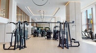 This premium fitness center is designed with both form and function in mind, offering a spacious and well-appointed environment for a complete workout experience. The architecture is defined by soaring double-height ceilings, tiered coffered detailing with integrated LED lighting, and warm-toned wood-framed glass partitions that lend the space an elegant, hotel-quality aesthetic.

The main workout floor is finished with light wood-toned flooring throughout, contributing to a clean and open ambiance. A dedicated cardio zone lines a wall of floor-to-ceiling windows draped with sheer white curtains, allowing generous natural light to fill the space while offering outdoor views — an ideal setting for treadmills, ellipticals, and stationary bikes. Additional cardio equipment, including air bikes and balance boards, is distributed across the open floor plan.

The strength training area is comprehensively equipped with a full array of cable machines, selectorized resistance stations, a power rack with a barbell bench setup, and a dedicated free weights section featuring color-coded dumbbells and wall mirrors. A functional training zone with turf flooring and suspension equipment adds versatility. A welcoming entrance foyer with an information kiosk and seating area completes the facility.