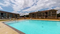 Located in Foley, Alabama, this community amenity space centers around a generously sized swimming pool with clear, turquoise water enclosed by a white metal fence. The pool deck is finished in neutral concrete, offering room for lounge chairs and umbrellas, making it well-suited for relaxation in the Alabama sun. A covered outdoor patio area extends alongside the pool enclosure, featuring shiplap-style siding in a muted sage green tone and white trim, consistent with the coastal-influenced architectural style of the broader community. This shaded patio is furnished with wicker sectional seating and coffee tables, creating a comfortable gathering space open to views of the surrounding grounds and multi-story residential buildings. Adjacent to the seating area, a poolside kitchenette station includes granite-topped counters, white cabinetry, bar seating, and a refrigerator, providing convenient amenities for entertaining. Multiple utility access doors are integrated into the structure. The entire space benefits from abundant natural light and open sky views, with well-maintained landscaping and ample parking visible in the background, contributing to an inviting, resort-inspired community atmosphere.