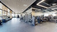 Located at 3170 W Olympic Blvd in Los Angeles, this sleek, single-floor fitness center at The Bora 3170 is designed for a comprehensive and modern workout experience. The space is defined by its clean architectural aesthetic, featuring a distinctive grid-panel ceiling with integrated linear LED lighting and track lighting throughout, all set above durable rubber flooring.

The main strength training area is generously proportioned, housing a full suite of professional-grade Technogym selectorized machines and free weights, with floor-to-ceiling mirrors amplifying the sense of space. A dedicated cardio zone lines the perimeter, where floor-to-ceiling glass windows flood the room with natural light and offer open views of the Los Angeles streetscape, creating an energizing workout environment.

A separate, glass-enclosed room provides a more intimate space for cycling and additional cardio equipment, while another enclosed zone features a fully equipped golf simulator with a turf hitting mat, projection screen, and digital tracking console — a standout amenity that sets this fitness center apart. Functional training equipment, including power racks, is also available throughout.

Supporting amenities include a water fountain and restroom facilities accessible directly from the gym floor, ensuring convenience for all users. The overall atmosphere is modern, well-lit, and professionally appointed.