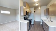 This single-floor residence offers a thoughtfully designed layout that balances functionality and modern aesthetics. The kitchen serves as a central highlight, featuring stainless steel appliances including a refrigerator, electric range, and microwave. Light wood-toned cabinetry extends to the upper walls, providing ample storage, while crisp white countertops offer generous prep space. The kitchen flooring is finished in durable wood-look planking that adds warmth and a contemporary feel. Flush-mount ceiling fixtures provide consistent, practical illumination throughout. The kitchen opens fluidly into an adjacent living area, which transitions to soft, neutral-toned carpeting, creating a comfortable and cohesive atmosphere. The layout is efficient and well-organized, with a clear sightline from the kitchen through the hallway toward additional rooms, suggesting a practical and connected floor plan. The overall color palette is calm and neutral, dominated by soft grays, warm wood tones, and white accents, contributing to a clean, modern ambiance throughout the space.