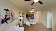 This single-floor residence offers a thoughtfully designed open-concept floor plan that integrates the living, dining, and kitchen areas into one cohesive and functional space. The living area is finished with plush carpeting and features crown molding and a ceiling fan, lending a comfortable and polished feel. A circular glass-top dining table with white chairs serves as the centerpiece of the dining area, positioned to flow naturally between the lounge and kitchen spaces. The kitchen is equipped with rich dark cabinetry, a black refrigerator, and a breakfast bar or counter-height surface that provides additional seating and workspace. Warm, neutral tones on the walls and ceilings create a cohesive and inviting ambiance throughout. Ceiling-mounted light fixtures provide consistent illumination across the open layout, complementing the natural light that filters in from the windows. A hallway visible at the rear of the space leads to additional rooms, including what appears to be a closet with bifold doors. Decorative touches such as a round wall mirror and framed wall art add personality and warmth to the interior. The overall design balances practicality with comfort, making it a welcoming and livable space.