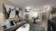 This elegantly designed single-floor residence offers a seamless open-concept layout that effortlessly connects the living, dining, and kitchen areas into one cohesive and inviting space. The living area is anchored by a plush gray sofa paired with a sculptural white ottoman, while large-scale abstract artwork adorns the walls, adding a bold artistic touch. A slender metal shelving unit serves as a subtle visual divider between the living and dining spaces, lending an airy sense of definition without disrupting the flow. The dining area features a classic round pedestal table surrounded by upholstered chairs, creating an intimate setting for meals. The kitchen is finished in crisp white cabinetry with sleek countertops, stainless steel appliances including a built-in microwave and refrigerator, and a peninsula that provides additional prep space and casual seating. Track lighting runs along the ceiling throughout, offering both functional illumination and a contemporary aesthetic. Crown molding adds a refined architectural detail, while wood-tone flooring grounds the space with warmth. The overall ambiance is sophisticated and modern, balancing clean lines with curated décor for a polished, welcoming atmosphere.