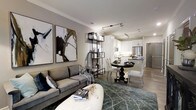 This elegantly designed single-floor residence offers a seamless open-concept layout that effortlessly connects the living, dining, and kitchen areas into one cohesive and inviting space. The living area is anchored by a plush gray sofa paired with a sculptural white ottoman, while large-scale abstract artwork adorns the walls, adding a bold artistic touch. A slender metal shelving unit serves as a subtle visual divider between the living and dining spaces, lending an airy sense of definition without disrupting the flow. The dining area features a classic round pedestal table surrounded by upholstered chairs, creating an intimate setting for meals. The kitchen is finished in crisp white cabinetry with sleek countertops, stainless steel appliances including a built-in microwave and refrigerator, and a peninsula that provides additional prep space and casual seating. Track lighting runs along the ceiling throughout, offering both functional illumination and a contemporary aesthetic. Crown molding adds a refined architectural detail, while wood-tone flooring grounds the space with warmth. The overall ambiance is sophisticated and modern, balancing clean lines with curated décor for a polished, welcoming atmosphere.