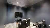 This single-floor screening room is thoughtfully designed to deliver an immersive cinematic experience. The space is finished with deep blue-gray walls that absorb light and minimize glare, creating a focused, theater-like atmosphere ideal for viewing. A large flat-screen television is mounted prominently on the front wall, serving as the centerpiece of the room, complemented by a media console positioned beneath it for equipment storage.

The seating arrangement features multiple plush leather recliner chairs with built-in cup holders, oriented directly toward the screen to maximize comfort and sightlines. The light wood-toned flooring provides a subtle contrast to the moody wall tones, while a drop ceiling with recessed lighting offers controlled, dimmable illumination that enhances the viewing ambiance without creating unwanted reflections.

The acoustic-friendly design of the room, with its enclosed layout and soft furnishings, contributes to a rich sound environment. Every element of this space — from the carefully chosen color palette to the purposeful furniture arrangement — has been curated to prioritize the viewing experience, making it a refined and functional retreat for entertainment and relaxation.