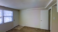 This two-floor space presents a straightforward and functional layout with a neutral color palette. The pictured room features beige-toned walls and wall-to-wall carpeting in a warm tan shade, creating a simple and understated ambiance. Two white doors are visible — one appearing to be an exterior entry door with a side window allowing in natural light, and the other an interior door likely leading to an adjacent room or closet. A subtle ceiling beam detail adds a minor architectural element to the otherwise smooth white ceiling. The room is clean and unfurnished, offering a clear view of the space's dimensions and potential. The overall aesthetic is modest and neutral, providing a blank canvas well-suited for personalization. With two floors, the property offers additional living or storage space beyond what is visible in this image. The consistent use of light, neutral tones throughout contributes to a bright and open feel, even in the absence of abundant windows in this particular area.