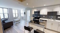 This single-floor space showcases a thoughtfully designed open-concept layout that seamlessly integrates the kitchen and living area. The kitchen is a standout feature, boasting white flat-panel cabinetry, a dark granite or quartz island with bar seating for two, and stainless steel appliances including a range, built-in microwave, and dishwasher. A slate-grey subway tile backsplash adds a modern contrast against the crisp white cabinetry and countertops. Light hardwood-style flooring flows continuously throughout the space, enhancing its clean, cohesive aesthetic. Recessed lighting provides consistent illumination across the open floor plan. The living area benefits from multiple windows fitted with horizontal blinds, allowing natural light to filter in while maintaining privacy. A white interior door leads to an adjacent room, suggesting additional private spaces beyond the main living area. The overall design palette — neutral whites, greys, and blacks — gives the space a contemporary, move-in-ready feel suited for modern living. The open layout maximizes the sense of space, making it feel airy and functional.