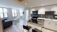 This single-floor space showcases a thoughtfully designed open-concept layout that seamlessly integrates the kitchen and living area. The kitchen is a standout feature, boasting white flat-panel cabinetry, a dark granite or quartz island with bar seating for two, and stainless steel appliances including a range, built-in microwave, and dishwasher. A slate-grey subway tile backsplash adds a modern contrast against the crisp white cabinetry and countertops. Light hardwood-style flooring flows continuously throughout the space, enhancing its clean, cohesive aesthetic. Recessed lighting provides consistent illumination across the open floor plan. The living area benefits from multiple windows fitted with horizontal blinds, allowing natural light to filter in while maintaining privacy. A white interior door leads to an adjacent room, suggesting additional private spaces beyond the main living area. The overall design palette — neutral whites, greys, and blacks — gives the space a contemporary, move-in-ready feel suited for modern living. The open layout maximizes the sense of space, making it feel airy and functional.