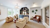 Located at 1540 Edgefield Dr in Garland, Texas, this single-floor living room is a well-appointed communal space that balances comfort and functionality with refined interior design. The room's most striking architectural feature is a dramatic arched built-in bookcase finished in a deep navy-gray, flanked by lower cabinetry that provides both display and storage. The unit is elegantly styled with books, greenery, and decorative accents, creating a warm focal point for the space. Neutral-toned carpeting runs throughout, complementing the crisp white walls and adding a sense of warmth underfoot. Seating is generously arranged, with cream upholstered armchairs and a rich leather accent chair gathered around a round wooden coffee table, encouraging conversation and relaxation. A drum pendant light fixture anchors the center of the room, while recessed lighting provides ambient illumination throughout. Decorative green resin wall pieces add an artistic touch to the walls. A dedicated workstation nook features a built-in white desk with dark cabinetry below, outfitted with a swing-arm wall sconce and set against a soft sage-green accent wall. Large windows invite ample natural light and offer views of the surrounding greenery, enhancing the open and airy atmosphere of this thoughtfully curated space.