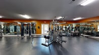 Located at 1000 Stonebrook Dr in Sanford, Florida, this single-floor fitness facility offers a comprehensive workout environment designed to accommodate a wide range of exercise needs. The main gym area is finished with durable dark rubber flooring and features bold orange accent walls paired with large wall-to-wall mirrors that enhance the sense of space and allow users to monitor their form during workouts. Overhead fluorescent lighting ensures the space is well-lit throughout.

The strength training section is well-appointed with a multi-station cable machine, weight benches, and a variety of selectorized resistance machines, providing options for both isolated and compound exercises. The cardio area includes multiple treadmills, stair climbers, and stationary bikes, several of which are equipped with individual display screens. Exercise balls, resistance equipment, and storage racks round out the functional fitness offerings.

An adjacent racquetball court is accessible through a glass door from the main gym floor. The court features a polished hardwood floor, high ceilings, and teal and white painted walls, with a viewing window that brings in natural light. This versatile space can also serve as a multi-purpose activity area. Together, the two areas form a well-rounded fitness facility suited for both individual training and active recreation.