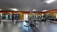 Located at 1000 Stonebrook Dr in Sanford, Florida, this single-floor fitness facility offers a comprehensive workout environment designed to accommodate a wide range of exercise needs. The main gym area is finished with durable dark rubber flooring and features bold orange accent walls paired with large wall-to-wall mirrors that enhance the sense of space and allow users to monitor their form during workouts. Overhead fluorescent lighting ensures the space is well-lit throughout.

The strength training section is well-appointed with a multi-station cable machine, weight benches, and a variety of selectorized resistance machines, providing options for both isolated and compound exercises. The cardio area includes multiple treadmills, stair climbers, and stationary bikes, several of which are equipped with individual display screens. Exercise balls, resistance equipment, and storage racks round out the functional fitness offerings.

An adjacent racquetball court is accessible through a glass door from the main gym floor. The court features a polished hardwood floor, high ceilings, and teal and white painted walls, with a viewing window that brings in natural light. This versatile space can also serve as a multi-purpose activity area. Together, the two areas form a well-rounded fitness facility suited for both individual training and active recreation.