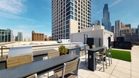 Located in the heart of Chicago's vibrant River North neighborhood, this rooftop amenity level at 717 N Wells St offers an impressive outdoor retreat with sweeping views of the city skyline. The space is designed for both relaxation and entertainment, combining functional amenities with an open-air atmosphere bathed in natural sunlight.

The rooftop terrace features a well-appointed outdoor kitchen and grilling station, complete with a built-in sink, stainless steel grill, and ample counter space finished in dark granite. Multiple seating arrangements — including high-top bar tables and chairs — provide comfortable spaces for dining and socializing against the dramatic Chicago skyline backdrop.

A stretch of lush artificial turf lawn adds a refreshing green element to the terrace, creating a casual lounge area that contrasts with the surrounding urban environment. Large planters with seasonal greenery and flowers are thoughtfully placed to enhance the aesthetic appeal of the space.

Interior access is provided through glass-and-metal framed doors with ADA-compliant features, including accessible restroom facilities and stairwell access clearly marked for safety and convenience. The transition between interior and exterior is seamless, with floor-to-ceiling glass offering unobstructed skyline views even from within. The overall design is modern and polished, reflecting the contemporary character of the surrounding River North district.