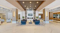 34 Eden River Park presents a thoughtfully designed single-floor space that combines an impressive arrival experience with functional, elegantly appointed areas. The grand entrance lobby sets the tone immediately, featuring large-format white marble-look tile flooring, soaring ceilings with a dramatic coffered detail finished in warm wood tones, and a pair of sculptural globe pendant chandeliers. A central seating area is anchored by a built-in fireplace flanked by decorative shelving, creating a welcoming focal point beneath the 34 Eden River Park branding. Floor-to-ceiling glass curtain walls flood the entry with abundant natural light and offer open views of the surrounding landscape.

Transitioning deeper into the space, herringbone-patterned hardwood flooring introduces a warmer, residential aesthetic. A comfortable lounge area features a slatted wood ceiling, a large wall-mounted display set against a matte gray feature wall, and built-in shelving accented by curated décor. Adjacent to this is a well-appointed leasing office with multiple workstations, gold pendant lighting, and ample glazing providing continuous outdoor views. A separate presentation area accommodates community displays and floor plan exhibits. Throughout, the design balances contemporary sophistication with approachable warmth, making the space both functional and visually engaging.