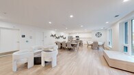 Located at 11 Wright Cres in Kingston, Ontario, this single-floor open-concept communal space offers a generous and well-appointed environment designed for both social gathering and relaxed lounging. The layout seamlessly integrates a fully equipped kitchen with a spacious living area, creating a cohesive and functional shared space.

The kitchen is outfitted with crisp white cabinetry, a white subway tile backsplash, stainless steel appliances including a refrigerator, stove, and microwave, and a large white island with bar seating for multiple guests. Colorful hexagonal wall art adds a playful accent to the otherwise clean, minimal aesthetic.

The living area is thoughtfully divided into distinct seating zones. A lounge section features a bold, tufted dark sofa paired with green armchairs, a large light wood coffee table, and contemporary artwork on the walls. A central conversation cluster with rounded white chairs surrounds a low circular table, while additional dining tables with upholstered chairs accommodate larger groups.

Throughout, light hardwood flooring, recessed ceiling lighting, and crisp white walls create a bright and airy ambiance. Structural columns add architectural character, and decorative touches such as a large wall clock and nature-inspired artwork contribute warmth and personality to this well-designed communal space.