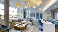 The Lounge is a thoughtfully designed communal space that combines modern aesthetics with a warm, inviting atmosphere. The room draws immediate attention upward with a striking cluster of woven, orb-shaped pendant lights suspended from the ceiling at varying heights, creating a sculptural focal point that doubles as ambient illumination throughout the space.

Large floor-to-ceiling windows line one wall, flooding the interior with abundant natural light and offering views of the surrounding greenery outside. The light hardwood flooring complements the room's cool, neutral color palette of whites, soft grays, and blues, lending a clean and contemporary feel.

The seating area anchors the space with a generous sectional sofa dressed in layered throw pillows in muted tones and pops of yellow and teal. A distinctive live-edge gold coffee table serves as a conversation piece, adding an organic, artistic element to the arrangement. A plush area rug grounds the seating zone comfortably.

To one side, a sleek bar-height counter with modern blue swivel stools offers an alternative spot for casual gathering. Colorful abstract artwork adorns the walls, contributing a curated, gallery-like quality. French doors lead to an adjacent space, enhancing the overall sense of openness and flow.