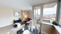 This single-floor residence offers an open-concept layout that seamlessly integrates the dining and living areas into one cohesive, functional space. The dining area features a dark-toned table set with place settings and woven placemats, positioned adjacent to large floor-to-ceiling sliding glass doors that lead to a private balcony. The balcony provides elevated panoramic views of the surrounding cityscape, filling the interior with natural light throughout the day.

The living area is carpeted and furnished with a dark sectional sofa accented by red and patterned cushions, complemented by a low-profile coffee table and a matching dark media console. Framed artwork on the neutral-toned walls adds a refined touch, while a floor lamp provides warm ambient lighting. A textured ceiling and visible HVAC vents reflect the practical, urban character of the space.

The overall ambiance is modern and urban, with a neutral color palette punctuated by bold accent tones. The connection between the interior living space and the outdoor balcony enhances the sense of openness, making city views a defining feature of this residence.