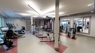 This fitness center offers a thoughtfully designed, single-floor layout that provides ample space for a full range of exercise activities. The open floor plan is anchored by a striking star-shaped ceiling light fixture that serves as both a functional and architectural focal point, casting bright, even illumination throughout the space. The flooring features a clean white base accented with bold red geometric insets, adding visual energy to the environment while clearly delineating workout zones. Structural columns are integrated seamlessly into the layout without impeding movement or equipment placement. The strength training area is well-stocked with a variety of resistance machines, free weight racks, and benches suited for both upper and lower body workouts. Large mirrors line one section of the wall, enhancing the sense of depth and allowing users to monitor their form during exercise. A secondary area, visible through an opening, provides additional equipment and benefits from natural light streaming in through windows. The overall ambiance is clean, modern, and functional, with neutral gray walls and a bright overhead lighting scheme that creates an energizing atmosphere well-suited for focused training sessions.