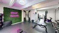 The Fitness Center at 372 Kettle Creek Road in Toms River, NJ, is a thoughtfully designed exercise facility that makes efficient use of its single-floor layout. The space is divided into distinct workout zones, each equipped to support a variety of training styles.

The main exercise room features bold black-and-white striped accent walls that lend a dynamic, energetic atmosphere to the space. A large wall-mounted flat-screen television sits above a full dumbbell rack with an accompanying weight bench, creating a dedicated free weights area. A tall mirror expands the sense of space and assists with form during training. A freestanding cable machine and additional adjustable benches provide options for strength training, while a green artificial moss wall displaying a neon "Mariners Cove" sign adds a distinctive branded touch and ambient lighting.

A separate alcove houses cardio equipment, including a stair climber, elliptical trainer, and stationary bicycle, all positioned near a window that allows natural light to filter in. The flooring throughout is durable rubber matting, appropriate for high-traffic fitness use. Overhead, circular modern light fixtures provide bright, even illumination across the facility.

A dedicated bathroom with tile flooring and neutral-toned walls rounds out the facility, offering convenience for users. The overall design balances functionality with a stylish, modern aesthetic.