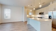 This well-designed 1-bedroom, 1-bathroom single-floor unit offers a clean, modern aesthetic throughout its efficiently organized layout. The open-concept kitchen and living area serves as the heart of the space, anchored by a sizeable central island with a crisp white countertop. The kitchen is outfitted with white shaker-style cabinetry, a white tile backsplash, stainless steel appliances including a refrigerator, microwave, and range, and globe pendant lights that hang above the island, adding a contemporary touch. Light wood-style flooring flows seamlessly throughout the entire unit, contributing to a bright and cohesive feel. A hallway connects the living area to the private bedroom, which features a ceiling fan, windows with blinds that allow natural light, and ample wall space. The full bathroom is finished with a single-sink vanity, white cabinetry, a framed mirror, and wood-style flooring. An open closet area with double hanging rods provides dedicated storage. The unit's consistent use of white walls, neutral tones, and modern fixtures creates a clean, move-in-ready atmosphere well-suited for comfortable everyday living.