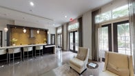 Located at 6215 Via La Cantera in San Antonio, Texas, this sophisticated single-floor clubhouse space offers a refined, contemporary design with a warm and welcoming ambiance. The open-plan layout is anchored by soaring ceilings and wide-plank hardwood flooring that flows seamlessly throughout, creating a sense of generous, airy space.

The main lounge area is appointed with elegant neutral-toned seating, including wingback chairs, a sleek sofa, and accent tables, all arranged around a decorative wall-mounted sunburst feature and an inset linear fireplace set against a smooth stone-clad wall. Floor-to-ceiling French doors line the exterior walls, flooding the interior with abundant natural light and offering lush views of tropical palms outside. Sheer drapery panels frame the windows, softening the light and adding an understated sense of luxury.

A standout kitchen and bar area features a dramatic gold-toned backsplash, dark cabinetry, a stainless steel range hood, and a broad white countertop island with modern bar seating — perfect for entertaining. Pendant lighting illuminates this space with a warm glow.

A secondary lounge area showcases an eye-catching lattice-patterned decorative wall panel, complemented by plush velvet seating arrangements ideal for intimate gatherings. Together, the varied seating zones and polished finishes create a cohesive, upscale environment suited for both social events and quiet relaxation.