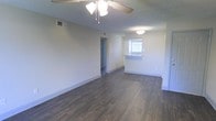 This single-floor space offers a clean, straightforward layout designed for comfortable everyday living. The main living area is open and airy, finished with dark hardwood-style flooring that runs throughout and provides a rich contrast against the neutral, light-toned walls. A ceiling fan with an integrated light fixture is centrally positioned overhead, complementing the textured ceiling and ensuring year-round comfort. Ventilation is supported by visible HVAC registers along the upper walls.

The living space flows naturally toward a kitchen area, which is distinguished by a raised breakfast bar — a practical feature that defines the boundary between the two zones while maintaining an open feel. A window above the kitchen sink allows natural light to filter into the space, brightening the interior. A flush-mount ceiling light in the kitchen area provides additional illumination.

A hallway visible from the main living area leads to additional rooms, including what appears to be a bathroom or closet space indicated by a door in a contrasting darker finish. White baseboards frame the perimeter of the rooms, adding a clean, finished look throughout. Overall, the space presents a simple, functional interior with consistent finishes.