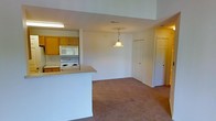 This single-floor residence offers a practical and comfortable open-concept layout that seamlessly connects the kitchen and living areas. The kitchen is equipped with white appliances, including an electric range and microwave, set against warm oak cabinetry that provides ample storage. A white peninsula counter extends into the space, creating a natural division between the kitchen and the adjoining living area while maintaining an open, airy feel. The living area is finished with wall-to-wall carpeting in a neutral tone, lending a warm and inviting atmosphere throughout. A decorative glass pendant light hangs centrally, serving as a charming focal point and providing warm ambient lighting to the space. The interior is painted in clean, neutral tones that enhance the sense of brightness and openness. Multiple doorways visible from the main area suggest additional rooms, including what appears to be a closet and further living quarters, adding to the overall functionality of the floor plan. The layout is straightforward and efficient, making the most of the available space while ensuring ease of movement throughout.