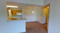 This single-floor residence offers a practical and comfortable open-concept layout that seamlessly connects the kitchen and living areas. The kitchen is equipped with white appliances, including an electric range and microwave, set against warm oak cabinetry that provides ample storage. A white peninsula counter extends into the space, creating a natural division between the kitchen and the adjoining living area while maintaining an open, airy feel. The living area is finished with wall-to-wall carpeting in a neutral tone, lending a warm and inviting atmosphere throughout. A decorative glass pendant light hangs centrally, serving as a charming focal point and providing warm ambient lighting to the space. The interior is painted in clean, neutral tones that enhance the sense of brightness and openness. Multiple doorways visible from the main area suggest additional rooms, including what appears to be a closet and further living quarters, adding to the overall functionality of the floor plan. The layout is straightforward and efficient, making the most of the available space while ensuring ease of movement throughout.