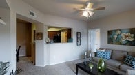 This single-floor residence offers a thoughtfully designed open-concept layout that promotes a seamless flow between living and kitchen areas. The living room is a welcoming space finished with plush carpeting and neutral-toned walls that create a bright, airy atmosphere. A ceiling fan with integrated lighting provides both comfort and illumination overhead. A defining architectural feature is the pass-through kitchen window, which connects the kitchen to the living area, enhancing the open feel while maintaining a defined cooking space. The kitchen features rich, dark cabinetry paired with a black refrigerator, creating a striking contrast against the lighter surroundings. The countertop ledge of the pass-through also serves as a convenient breakfast bar or display area. Natural light filters throughout the space, contributing to its inviting ambiance. A doorway visible from the living room leads to additional rooms, suggesting a well-organized floor plan with private sleeping quarters. Clean white trim and doors add a crisp, polished finish throughout. Overall, the residence balances functionality with a comfortable, casual aesthetic well-suited for everyday living.