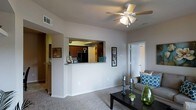 This single-floor residence offers a thoughtfully designed open-concept layout that promotes a seamless flow between living and kitchen areas. The living room is a welcoming space finished with plush carpeting and neutral-toned walls that create a bright, airy atmosphere. A ceiling fan with integrated lighting provides both comfort and illumination overhead. A defining architectural feature is the pass-through kitchen window, which connects the kitchen to the living area, enhancing the open feel while maintaining a defined cooking space. The kitchen features rich, dark cabinetry paired with a black refrigerator, creating a striking contrast against the lighter surroundings. The countertop ledge of the pass-through also serves as a convenient breakfast bar or display area. Natural light filters throughout the space, contributing to its inviting ambiance. A doorway visible from the living room leads to additional rooms, suggesting a well-organized floor plan with private sleeping quarters. Clean white trim and doors add a crisp, polished finish throughout. Overall, the residence balances functionality with a comfortable, casual aesthetic well-suited for everyday living.
