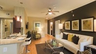This single-floor residence showcases an open-concept layout that seamlessly connects the living and dining areas. The living room serves as the focal point, anchored by a bold dark accent wall that contrasts elegantly with the warm beige tones found throughout the rest of the space. A white sofa adorned with earth-toned and green accent pillows sits opposite a sleek glass-top coffee table, creating a polished yet inviting seating area. Framed artwork on the accent wall and a pair of wall-mounted sconces add decorative character.

A ceiling fan with an integrated light fixture provides both airflow and ambient lighting overhead. Light hardwood flooring extends throughout, lending warmth and continuity to the space. The dining area, positioned near a pendant-lit bar counter, features modern white bar-style seating, offering a casual dining option. A cozy accent chair with a floor lamp occupies a corner near a window, creating a comfortable reading nook. Hallways lead to additional rooms, including what appears to be a bathroom and bedroom, completing this compact yet thoughtfully designed layout.