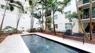 This inviting outdoor courtyard space offers a tranquil escape defined by a clean-lined lap pool as its centerpiece. The pool features elegant fountain jets that add gentle movement and ambiance to the water's surface, surrounded by a smooth stone tile deck that provides ample space for relaxation. The courtyard is enclosed by a multi-story modern building with a crisp white stucco exterior, large windows, and private balconies that overlook the lush interior grounds. Towering royal palms and slender bamboo rise dramatically throughout the space, creating a tropical canopy that filters natural light and lends a resort-like atmosphere. Dense tropical plantings, including shrubs and ground cover set in rich mulched beds, fill the surrounding landscape with vibrant greenery. A classic bench positioned along the pool deck invites residents to sit and enjoy the peaceful environment. The overall ambiance is one of refined tropical living — a well-maintained, private oasis that balances modern architectural design with lush, verdant landscaping to create a calm and visually striking communal outdoor experience.